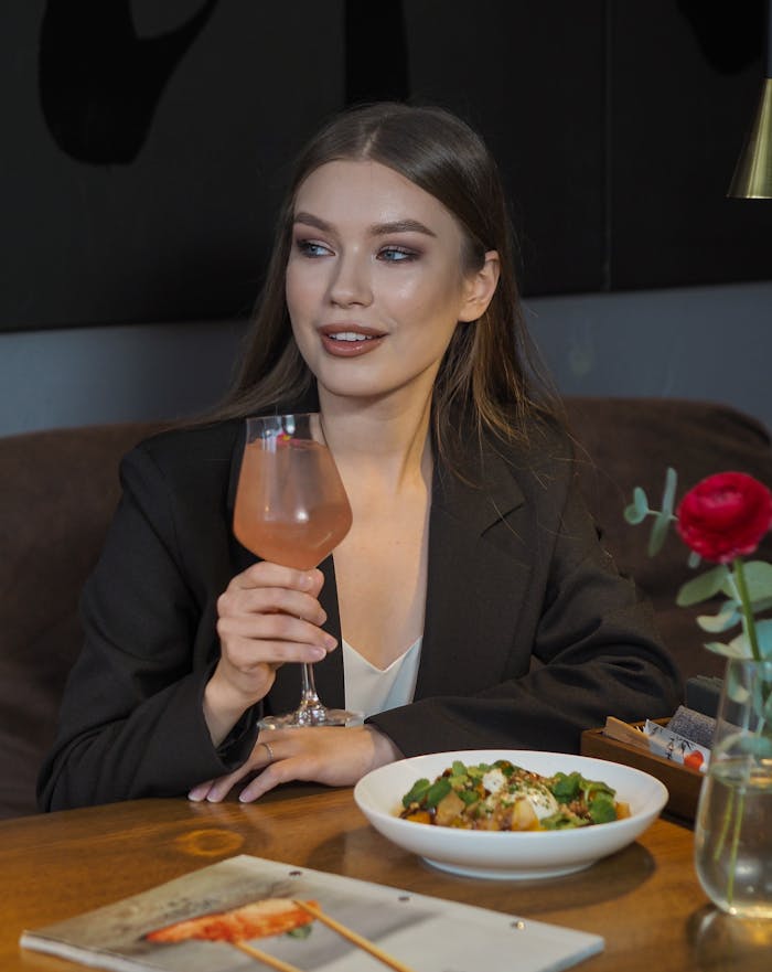 Elegant woman in a suit holding a cocktail, seated at a restaurant with food and decor.