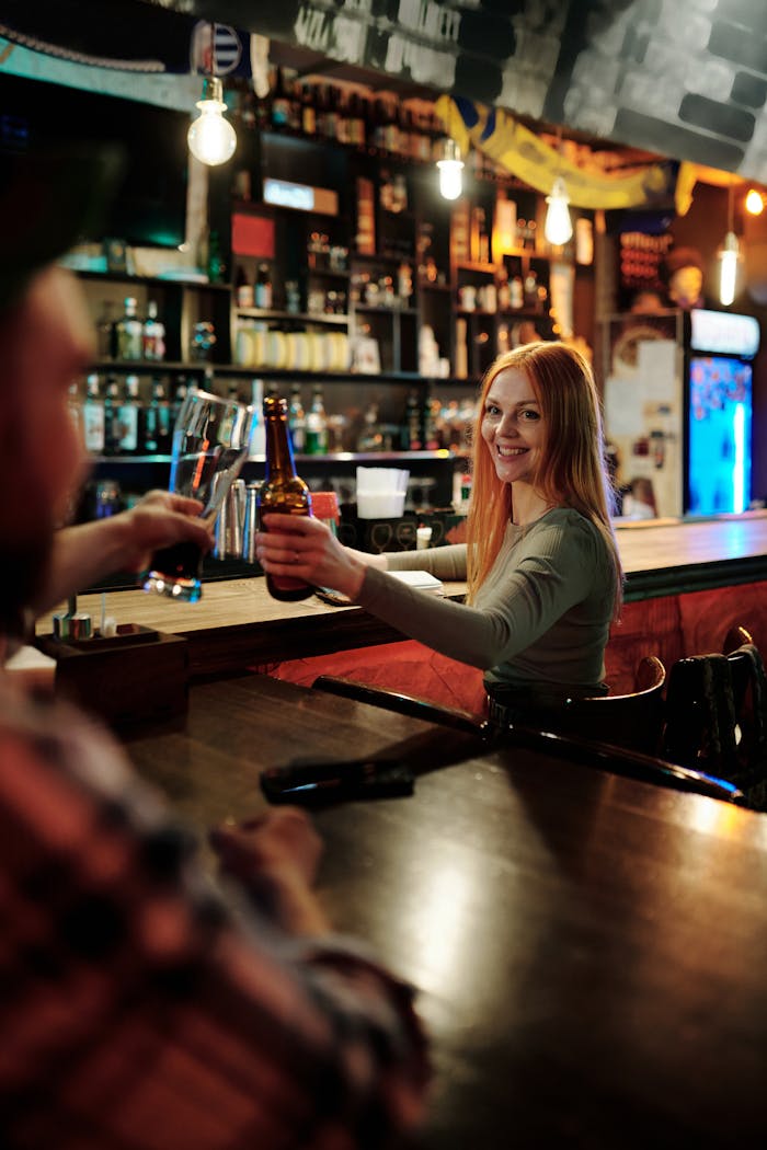 A cheerful young woman raises a beer bottle in a lively bar, embodying social enjoyment and night out vibes.