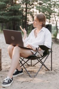 A woman sitting on a folding chair using a laptop outdoors, showcasing remote work in nature.