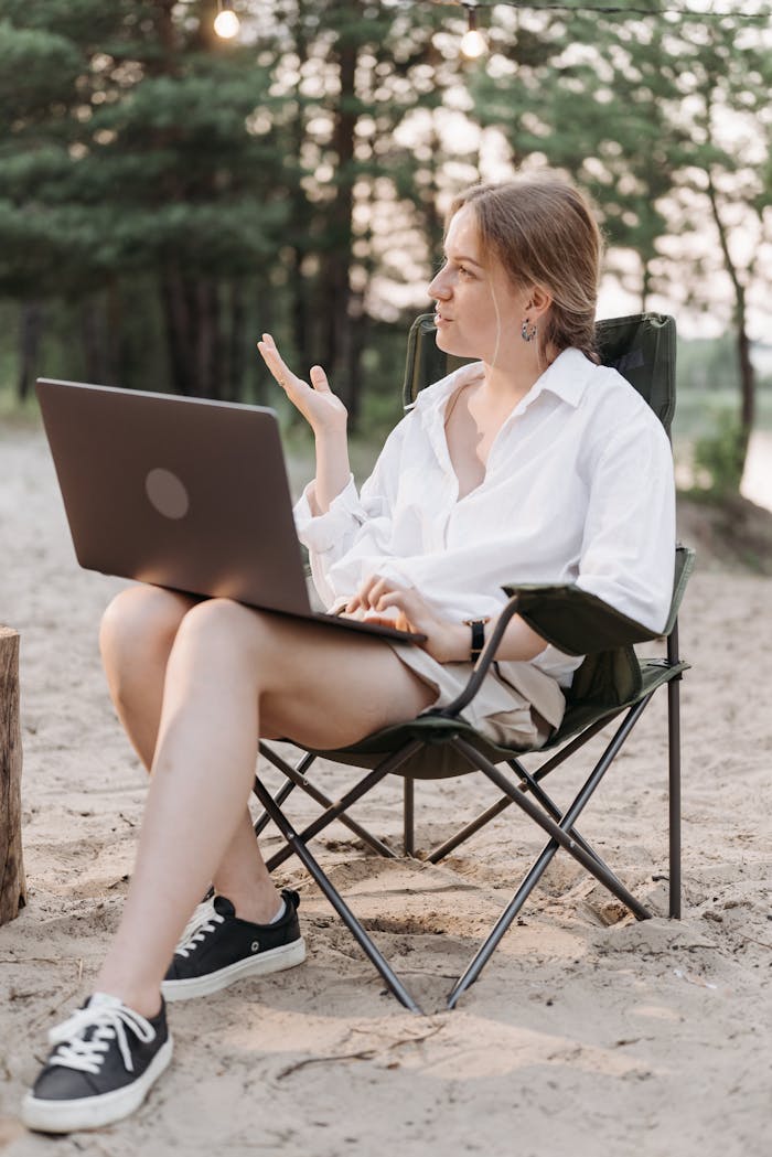 A woman sitting on a folding chair using a laptop outdoors, showcasing remote work in nature.